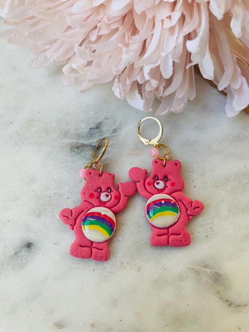 May include: A pair of pink Care Bear earrings with rainbow belly buttons. The earrings are made of polymer clay and have gold hoops.