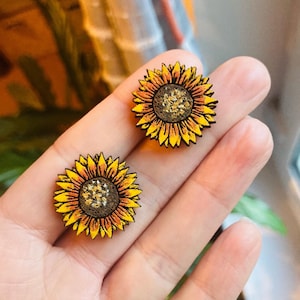 Puede incluir: Un par de pendientes de girasol de madera con pétalos amarillos, centros marrones y detalles de purpurina dorada.