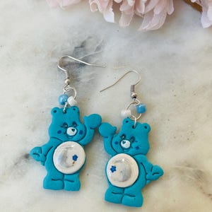 May include: A pair of blue Care Bear earrings with a white moon and stars design. The earrings are made of polymer clay and have silver hooks.