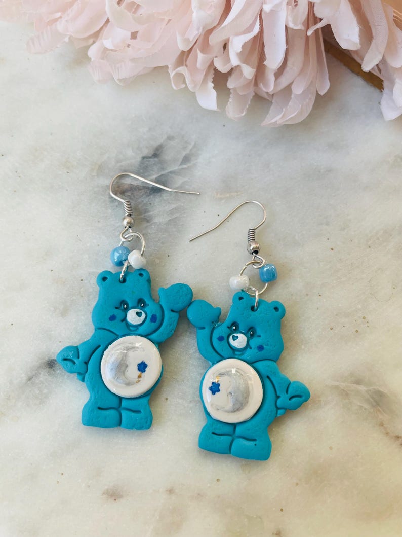 May include: A pair of blue Care Bear earrings with a white moon and stars design. The earrings are made of polymer clay and have silver hooks.
