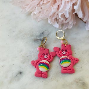 May include: Pink Care Bear earrings with rainbow belly buttons and gold hoops.