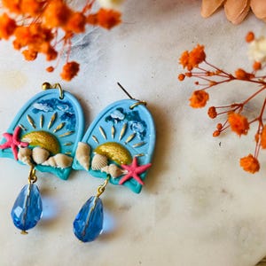 May include: A pair of arch-shaped earrings featuring a light blue sky, a gold sun, white clouds, small seashells, and a pink starfish. Each earring hangs from a gold post and includes a faceted, teardrop-shaped blue crystal bead dangling below.