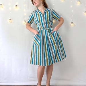 May include: A knee-length dress featuring a vertical striped pattern in shades of teal, olive green, white, and grey. The garment includes a collared neckline, short sleeves, a button-down front, a matching waist tie, and two large front pockets.