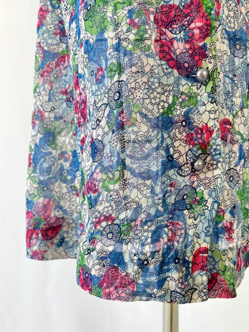 Close up of the hem A vintage 1940's floral print shirtwaist dress with short sleeves and a button-down front. The dress features a collared neckline and a midi-length skirt. The fabric is semi-sheer and has a blue, red, and green floral pattern that resembles stained glass. The dress is displayed on a mannequin against a white backdrop.