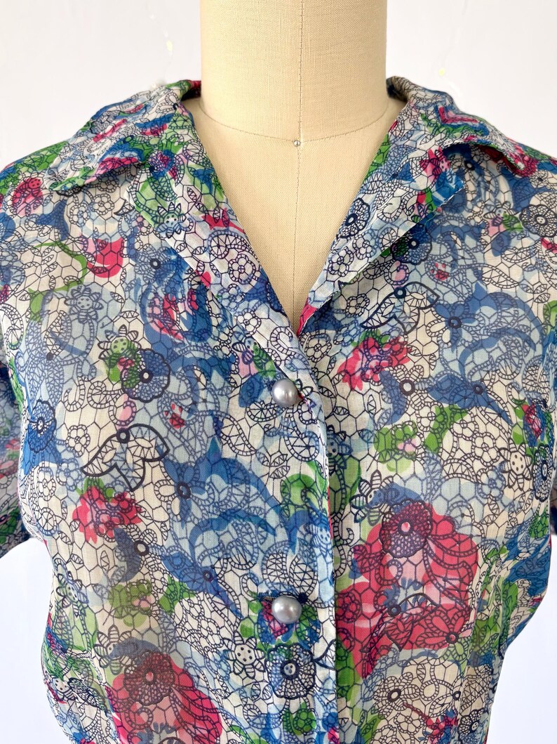 Close up of the neckline A vintage 1940's floral print shirtwaist dress with short sleeves and a button-down front. The dress features a collared neckline and a midi-length skirt. The fabric is semi-sheer and has a blue, red, and green floral pattern that resembles stained glass. The dress is displayed on a mannequin against a white backdrop.