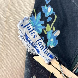 Może przedstawiać: Czarna odzież z niebiesko-zielonym wzorem kwiatowym. Dołączona jest biała metka z napisem Jules Canton FROCKS GUARANTEED WASHABLE. Metka jest częściowo przytrzymywana drewnianą klamerką do bielizny i ma postrzępione krawędzie.