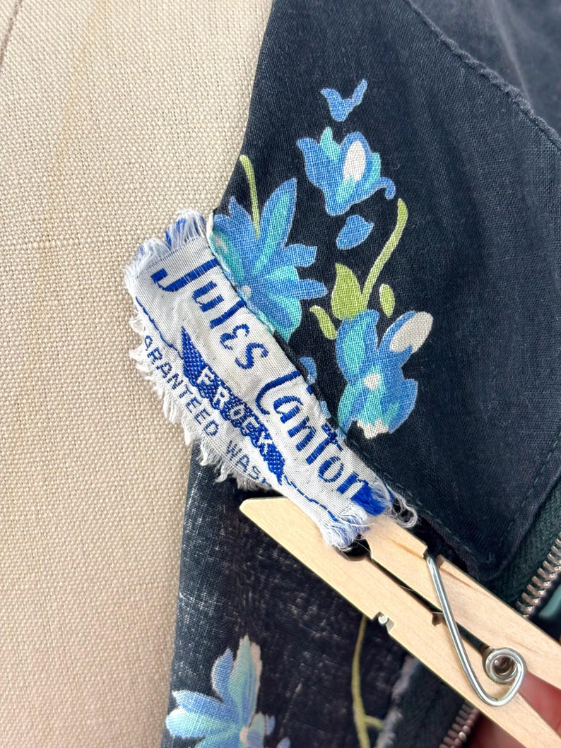 Może przedstawiać: Czarna odzież z niebiesko-zielonym wzorem kwiatowym. Dołączona jest biała metka z napisem Jules Canton FROCKS GUARANTEED WASHABLE. Metka jest częściowo przytrzymywana drewnianą klamerką do bielizny i ma postrzępione krawędzie.