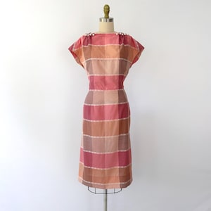 May include: A vintage, knee-length dress in a plaid pattern of pink, peach, and brown hues. The dress features short sleeves, a fitted waist, and a row of small, white pearl-like embellishments at the shoulders. The dress is displayed on a mannequin.