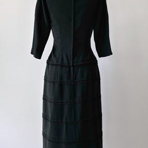 Back view: Authentic 1950s black wiggle dress with three-quarter sleeves and an hourglass silhouette. The dress features a row of small buttons down the front and decorative lace trim along the neckline and skirt. The dress is displayed on a size 2 mannequin.
