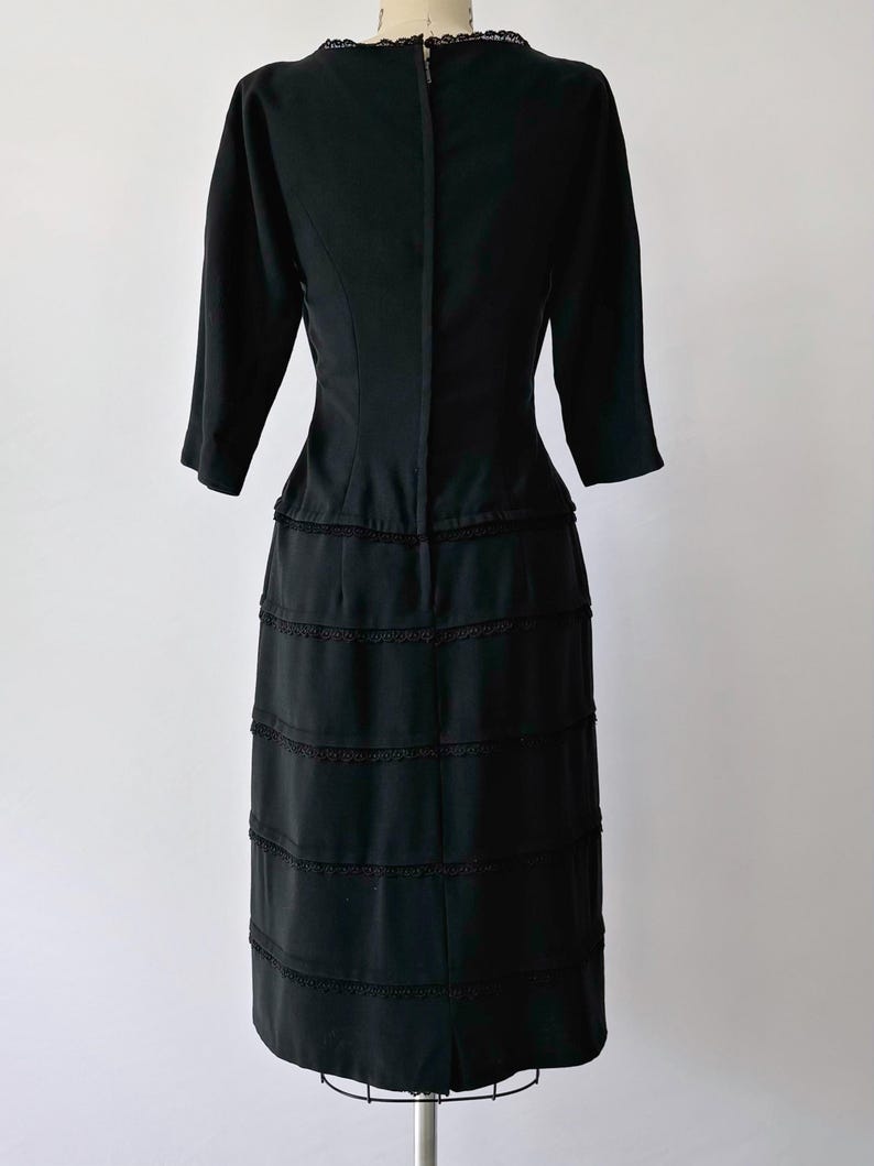 Back view: Authentic 1950s black wiggle dress with three-quarter sleeves and an hourglass silhouette. The dress features a row of small buttons down the front and decorative lace trim along the neckline and skirt. The dress is displayed on a size 2 mannequin.