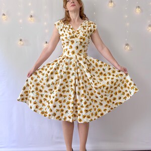 May include: A knee-length dress featuring a white base with an all-over sunflower print. The garment has a V-neckline, cap sleeves, and a fitted bodice that transitions into a pleated, flared skirt. The fabric displays yellow and brown flowers.