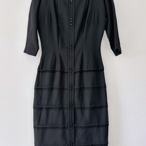 Vintage 1950s inky black wiggle dress shown hanging on a black velvet hanger and white background. It has a fitted bodice and a fitted knee-length skirt.