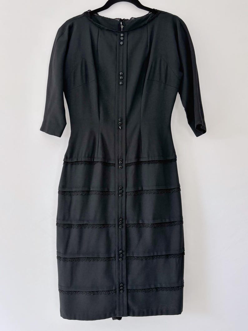 Vintage 1950s inky black wiggle dress shown hanging on a black velvet hanger and white background. It has a fitted bodice and a fitted knee-length skirt.