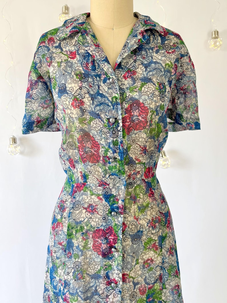 Close up of the bodice A vintage 1940's floral print shirtwaist dress with short sleeves and a button-down front. The dress features a collared neckline and a midi-length skirt. The fabric is semi-sheer and has a blue, red, and green floral pattern that resembles stained glass. The dress is displayed on a mannequin against a white backdrop.