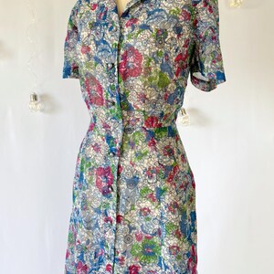 Close up of the bodice. A vintage 1940's floral print shirtwaist dress with short sleeves and a button-down front. The dress features a collared neckline and a midi-length skirt. The fabric is semi-sheer and has a blue, red, and green floral pattern that resembles stained glass. The dress is displayed on a mannequin against a white backdrop.