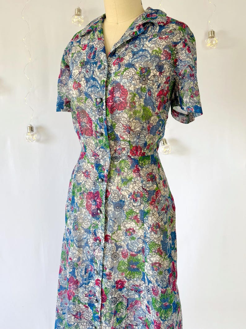 Close up of the bodice. A vintage 1940's floral print shirtwaist dress with short sleeves and a button-down front. The dress features a collared neckline and a midi-length skirt. The fabric is semi-sheer and has a blue, red, and green floral pattern that resembles stained glass. The dress is displayed on a mannequin against a white backdrop.