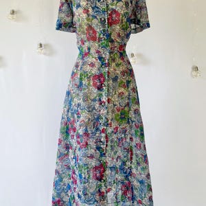 A vintage 1940's floral print shirtwaist dress with short sleeves and a button-down front. The dress features a collared neckline and a midi-length skirt. The fabric is semi-sheer and has a blue, red, and green floral pattern that resembles stained glass. The dress is displayed on a mannequin against a white backdrop.