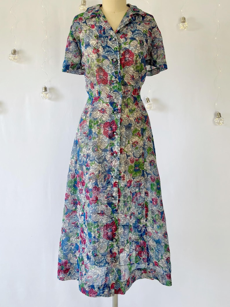 A vintage 1940's floral print shirtwaist dress with short sleeves and a button-down front. The dress features a collared neckline and a midi-length skirt. The fabric is semi-sheer and has a blue, red, and green floral pattern that resembles stained glass. The dress is displayed on a mannequin against a white backdrop.