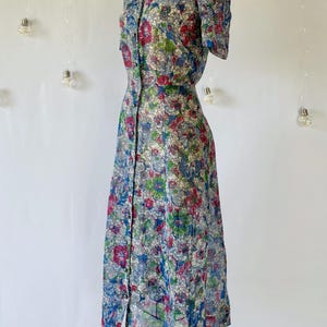From the left. A vintage 1940's floral print shirtwaist dress with short sleeves and a button-down front. The dress features a collared neckline and a midi-length skirt. The fabric is semi-sheer and has a blue, red, and green floral pattern that resembles stained glass. The dress is displayed on a mannequin against a white backdrop.