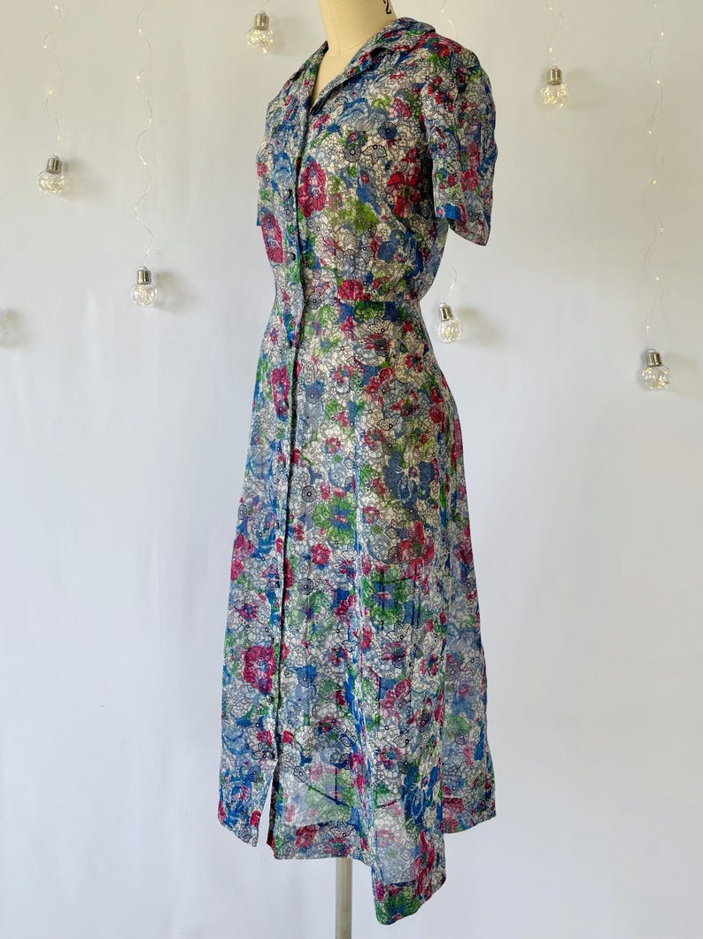 From the left. A vintage 1940's floral print shirtwaist dress with short sleeves and a button-down front. The dress features a collared neckline and a midi-length skirt. The fabric is semi-sheer and has a blue, red, and green floral pattern that resembles stained glass. The dress is displayed on a mannequin against a white backdrop.