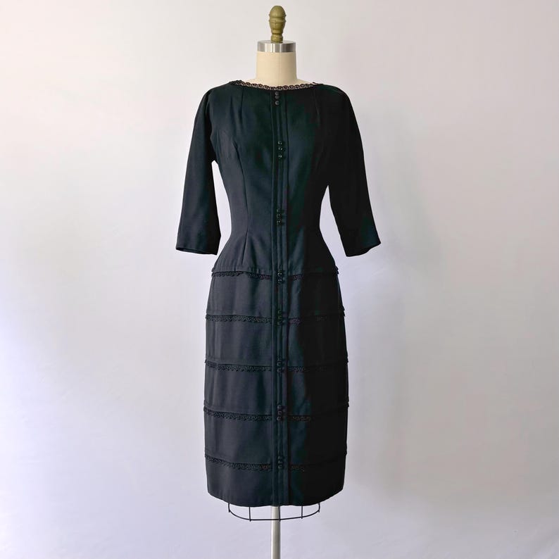 Authentic 1950s inky black wiggle dress with three-quarter sleeves and an hourglass silhouette. The dress features a row of small buttons down the front and decorative lace trim along the neckline and skirt. The dress is displayed on a size 2 mannequin.