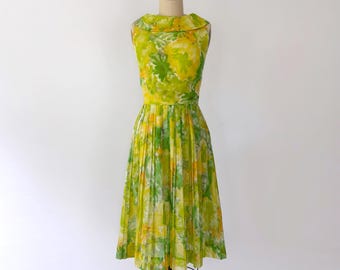 Può includere: Un abito vintage senza maniche con un motivo floreale nei toni del giallo e del verde. L'abito presenta una gonna a pieghe e uno scollo tondo. L'abito è esposto su un manichino su sfondo bianco.