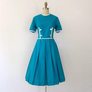 Puede incluir: Vestido turquesa vintage con falda plisada y mangas cortas. El vestido presenta ribetes blancos en el cuello, las mangas y la cintura, con botones blancos y una línea de acento blanca. El vestido se exhibe en un maniquí.