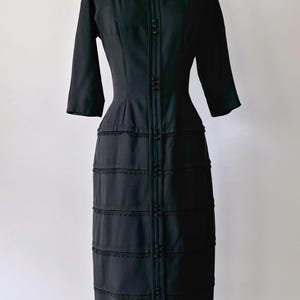 Authentic 1950s black wiggle dress with three-quarter sleeves and an hourglass silhouette. The dress features a row of small buttons down the front and decorative lace trim along the neckline and skirt. The dress is displayed on a size 2 mannequin.