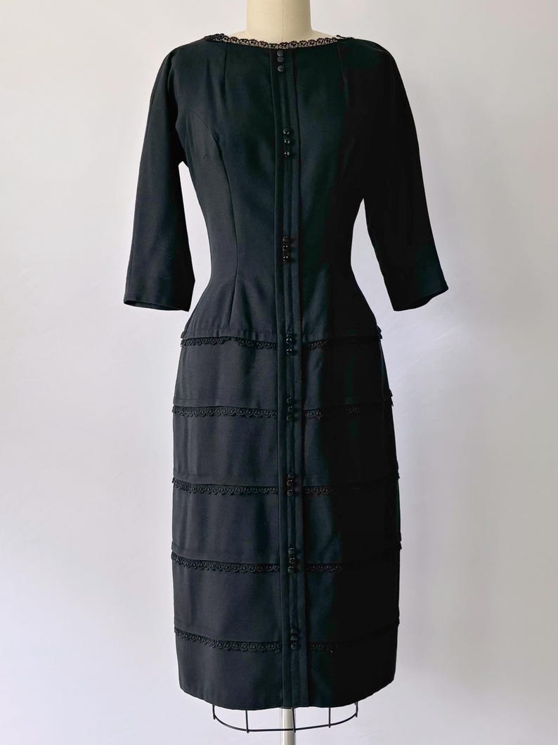 Authentic 1950s black wiggle dress with three-quarter sleeves and an hourglass silhouette. The dress features a row of small buttons down the front and decorative lace trim along the neckline and skirt. The dress is displayed on a size 2 mannequin.
