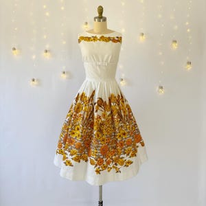 May include: A vintage white dress with a floral pattern in shades of yellow and brown. The dress features a fitted bodice and a flared skirt, displayed on a mannequin. The dress has a scoop neckline and is knee-length.