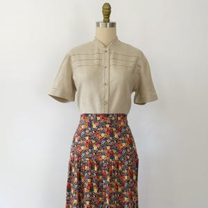 May include: A vintage two-piece outfit displayed on a mannequin. The top is a tan short-sleeved shirt with a mandarin collar and decorative horizontal stitching. The skirt is a high-waisted, floral print midi skirt with a pleated design. The floral pattern includes red, yellow, and blue flowers.