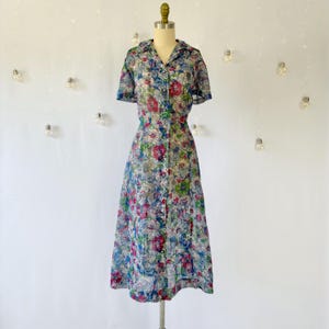 A vintage 1940's floral print shirtwaist dress with short sleeves and a button-down front. The dress features a collared neckline and a midi-length skirt. The fabric is semi-sheer and has a blue, red, and green floral pattern that resembles stained glass. The dress is displayed on a mannequin against a white backdrop.