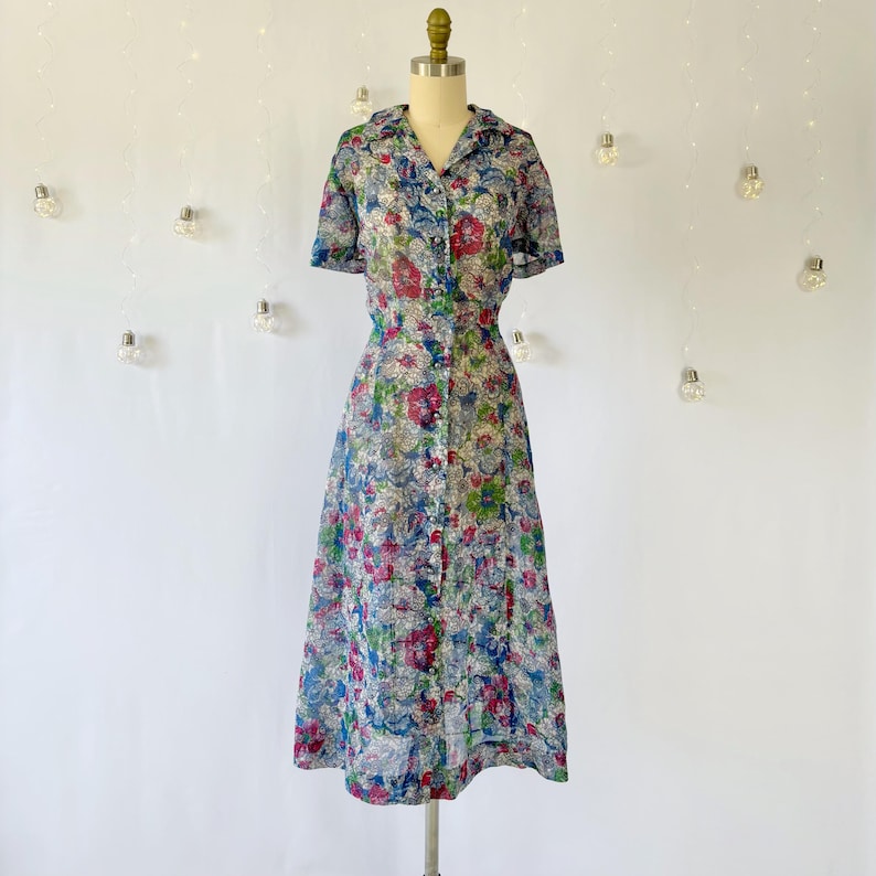 A vintage 1940's floral print shirtwaist dress with short sleeves and a button-down front. The dress features a collared neckline and a midi-length skirt. The fabric is semi-sheer and has a blue, red, and green floral pattern that resembles stained glass. The dress is displayed on a mannequin against a white backdrop.