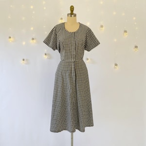 May include: A black and white checkered dress with short sleeves and a midi length. The dress is displayed on a mannequin against a white background with string lights.