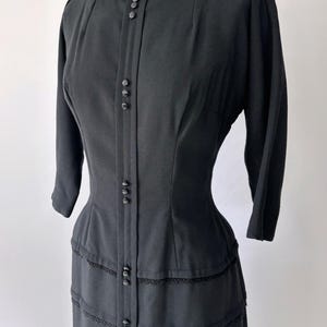 Close-up of the bodice at an angle of the 1950s black wiggle dress. It has a boat neck, princess seams, and three-quarter sleeves. There are groups of small decorative black buttons down the center vertical of the dress.