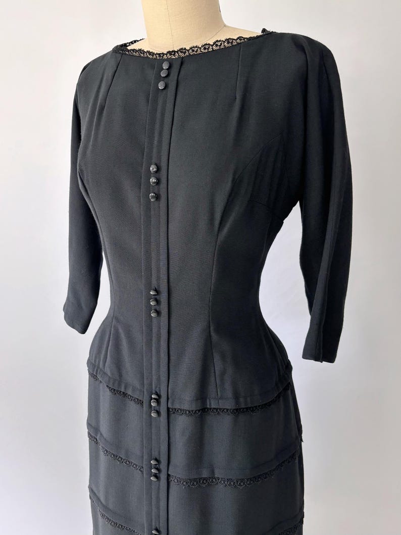 Close-up of the bodice at an angle of the 1950s black wiggle dress. It has a boat neck, princess seams, and three-quarter sleeves. There are groups of small decorative black buttons down the center vertical of the dress.
