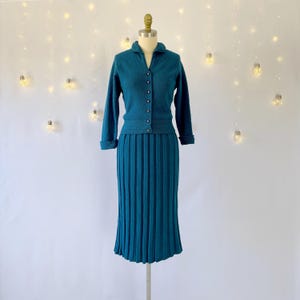 May include: Teal-colored vintage knitwear set, consisting of a collared cardigan with button closures and a matching pleated midi skirt. The set is displayed on a mannequin against a white backdrop with string lights. The cardigan has long sleeves with rolled cuffs.