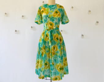 Può includere: Un abito floreale vintage nei toni del turchese, giallo e verde. L'abito presenta uno scollo tondo, maniche corte e una vita aderente con un fiocco abbinato. La gonna è ampia e arriva a metà polpaccio. L'abito è esposto su un manichino.
