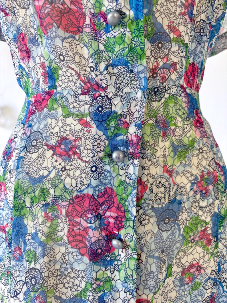 Close up of the waist A vintage 1940's floral print shirtwaist dress with short sleeves and a button-down front. The dress features a collared neckline and a midi-length skirt. The fabric is semi-sheer and has a blue, red, and green floral pattern that resembles stained glass. The dress is displayed on a mannequin against a white backdrop.