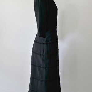 Side view: Authentic 1950s black wiggle dress with three-quarter sleeves and an hourglass silhouette. The dress features a row of small buttons down the front and decorative lace trim along the neckline and skirt. The dress is displayed on a size 2 mannequin.