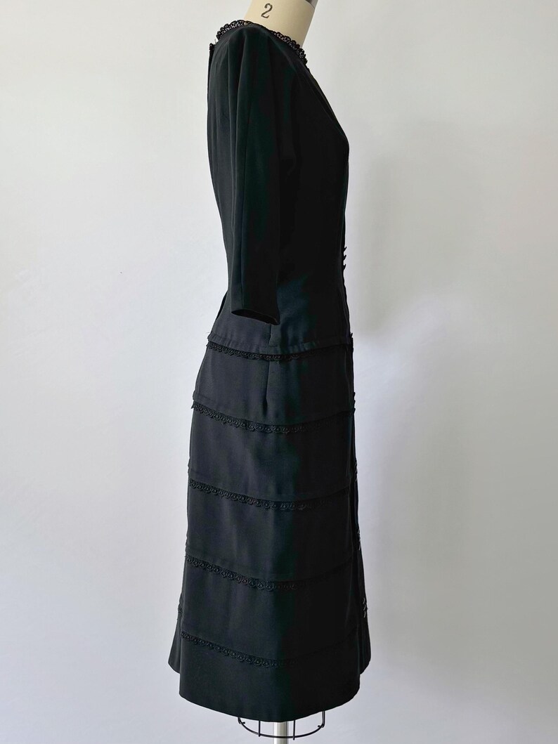 Side view: Authentic 1950s black wiggle dress with three-quarter sleeves and an hourglass silhouette. The dress features a row of small buttons down the front and decorative lace trim along the neckline and skirt. The dress is displayed on a size 2 mannequin.