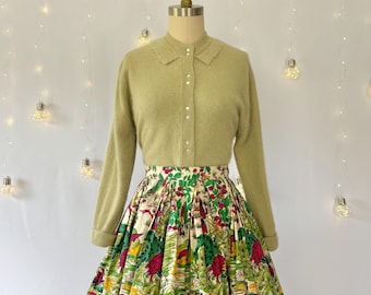 Può includere: Un cardigan a maniche lunghe color verde oliva chiaro con abbottonatura, abbinato a una gonna a vita alta con una vivace stampa botanica e di uccelli multicolore, con sfumature di verde, rosso, giallo e bianco su fondo color crema.