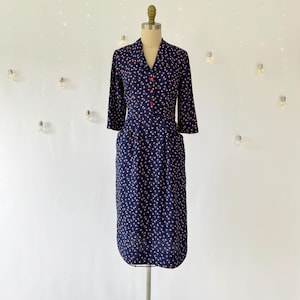 May include: A navy blue dress with a small floral pattern in pink and white. The dress has a V-neck, three-quarter sleeves, and red buttons down the front. It features two pockets and is displayed on a mannequin.