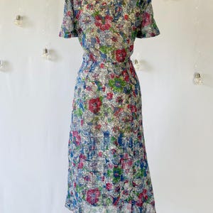 From behind. A vintage 1940's floral print shirtwaist dress with short sleeves and a button-down front. The dress features a collared neckline and a midi-length skirt. The fabric is semi-sheer and has a blue, red, and green floral pattern that resembles stained glass. The dress is displayed on a mannequin against a white backdrop.