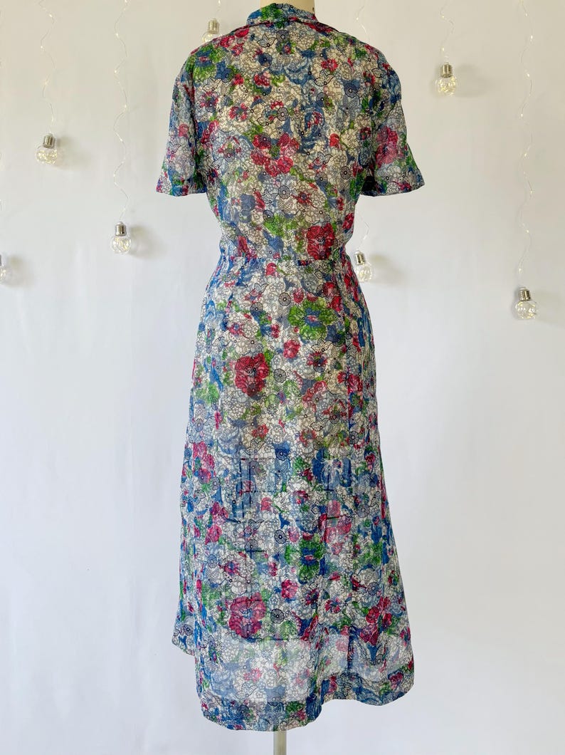 From behind. A vintage 1940's floral print shirtwaist dress with short sleeves and a button-down front. The dress features a collared neckline and a midi-length skirt. The fabric is semi-sheer and has a blue, red, and green floral pattern that resembles stained glass. The dress is displayed on a mannequin against a white backdrop.