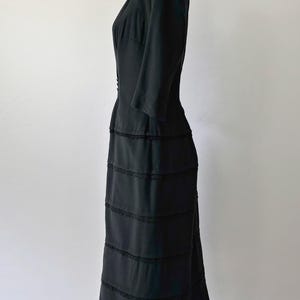 Side view: Authentic 1950s black wiggle dress with three-quarter sleeves and an hourglass silhouette. The dress features a row of small buttons down the front and decorative lace trim along the neckline and skirt. The dress is displayed on a size 2 mannequin.