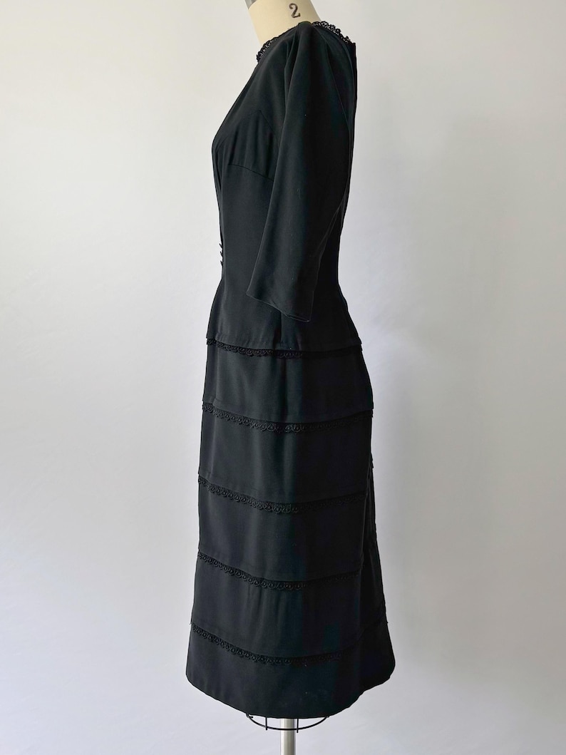 Side view: Authentic 1950s black wiggle dress with three-quarter sleeves and an hourglass silhouette. The dress features a row of small buttons down the front and decorative lace trim along the neckline and skirt. The dress is displayed on a size 2 mannequin.