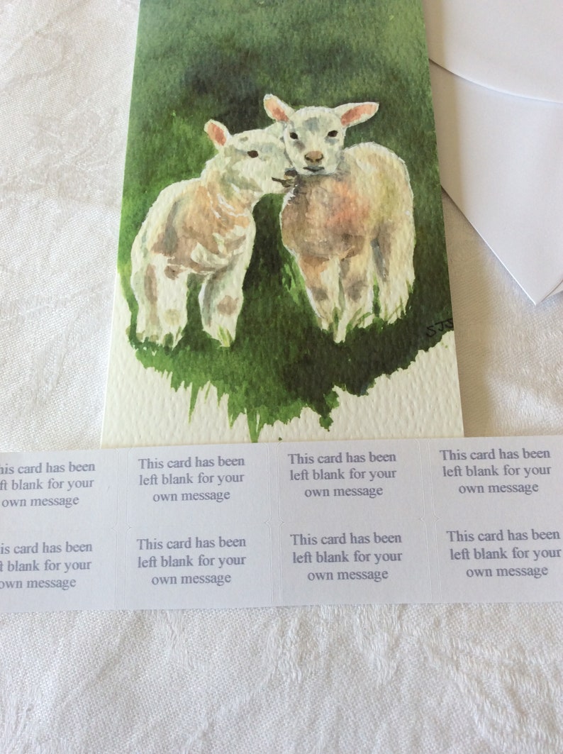 May include: A watercolor painting of two white lambs standing in a green field. The card has been left blank for a message.