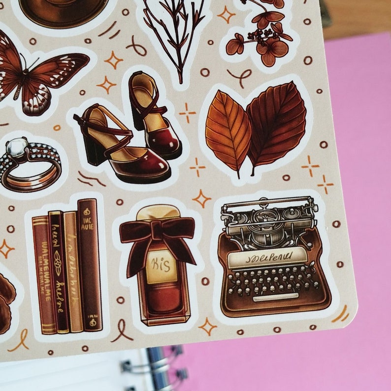 May include: A set of vintage-style stickers featuring a typewriter, a bottle of perfume, a pair of shoes, a butterfly, a ring, a stack of books, and leaves. The stickers are on a beige background with white outlines.
