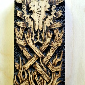 May include: A rectangular wooden carving with a detailed design. The central image is a stylized skull with antlers, surrounded by interwoven branches and flame-like shapes. The wood has a dark, textured background and lighter, detailed carvings.